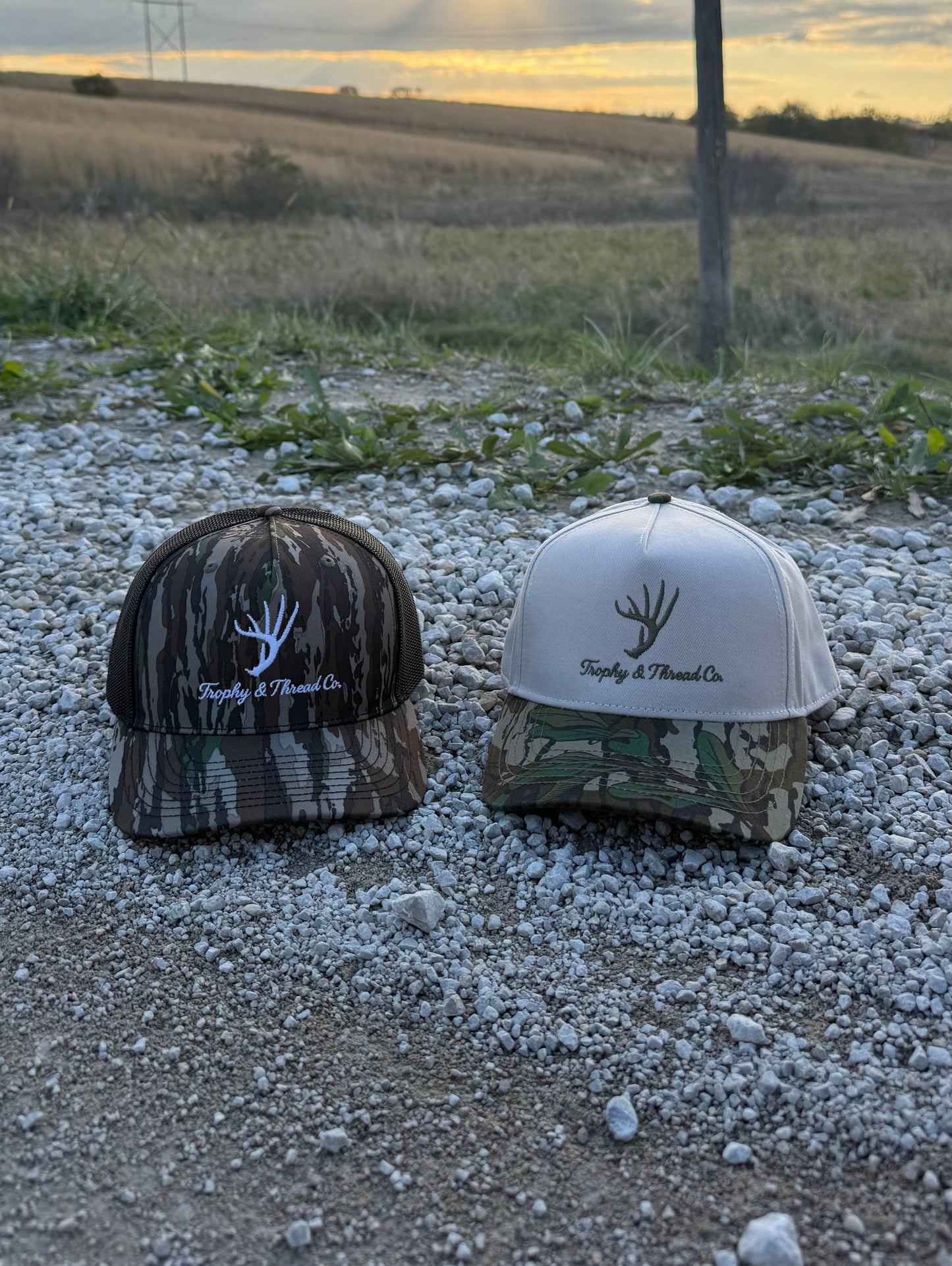 Whitetail Deer Hat Trophy and Thread Co Two caps, one camouflage and one white, on a gravel surface with a field and sunset in the background. 