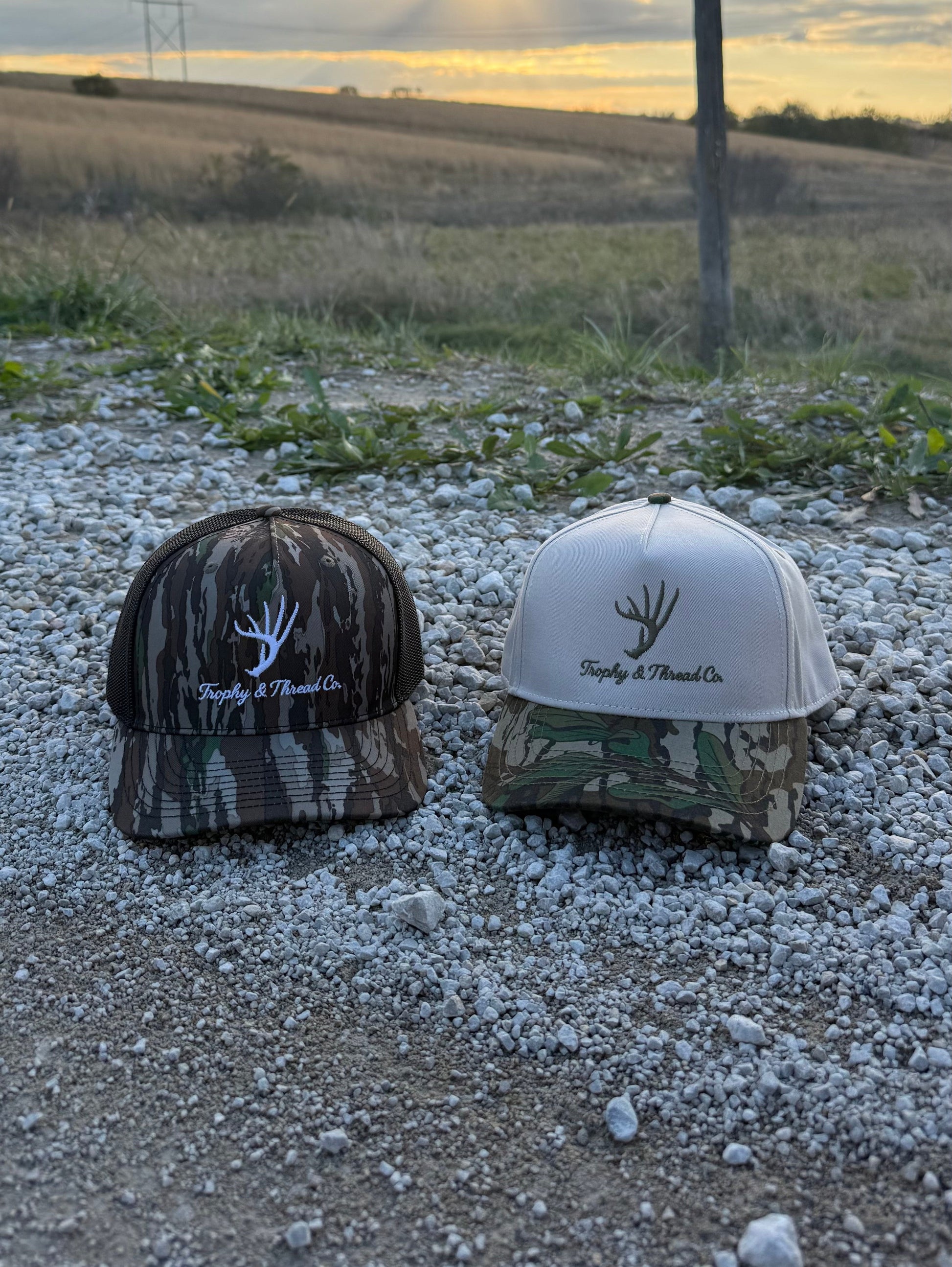 Whitetail Deer Hat Trophy and Thread Co Two caps, one camouflage and one white, on a gravel surface with a field and sunset in the background. 
