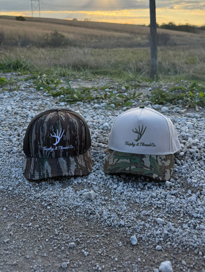 Whitetail Deer Hat Trophy and Thread Co Two caps, one camouflage and one white, on a gravel surface with a field and sunset in the background. 